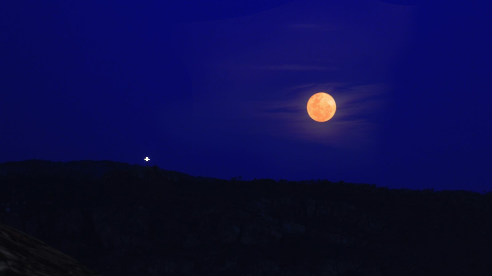 Lua cheia laranja subindo acima de uma serra escura em Diamantina, Minas Gerais; a Lua parece maior no horizonte por ilusão