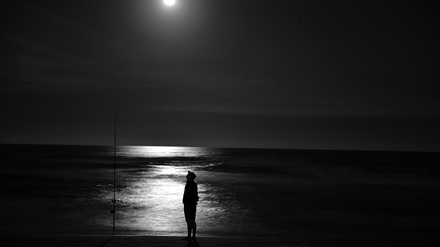Pescador em silhueta sob lua cheia numa praia à noite, tentando fisgar peixe