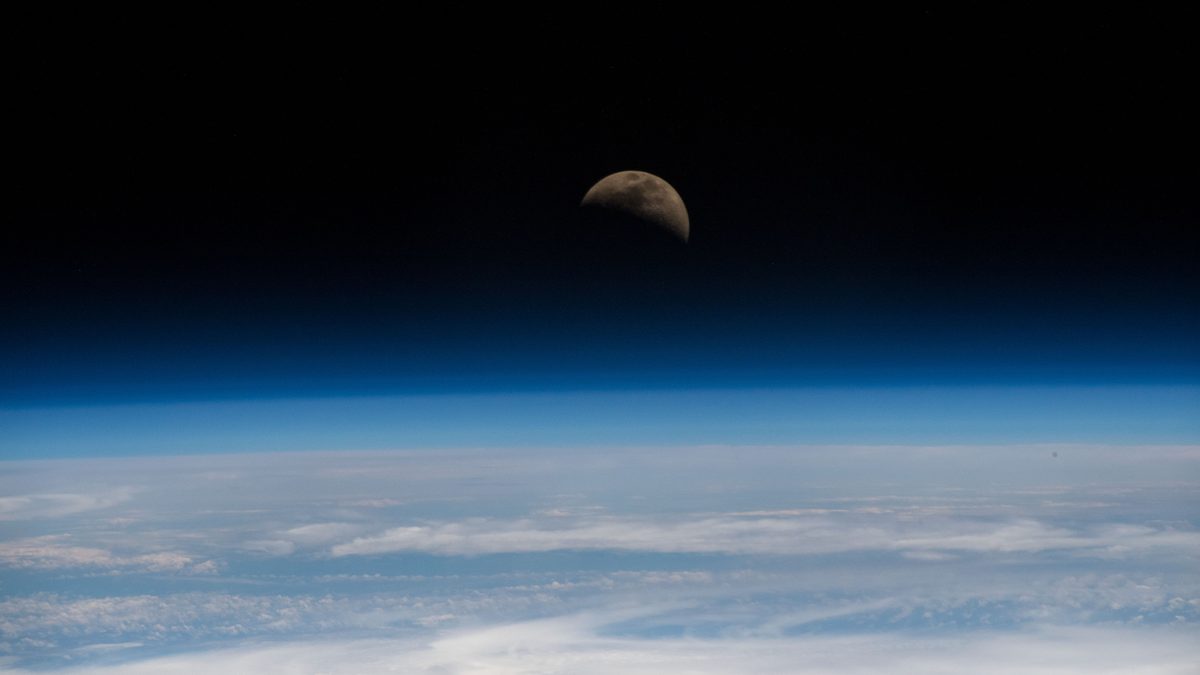 Lua em quarto crescente, com metade do disco iluminado, acima da curva azul da atmosfera da Terra e de nuvens brancas