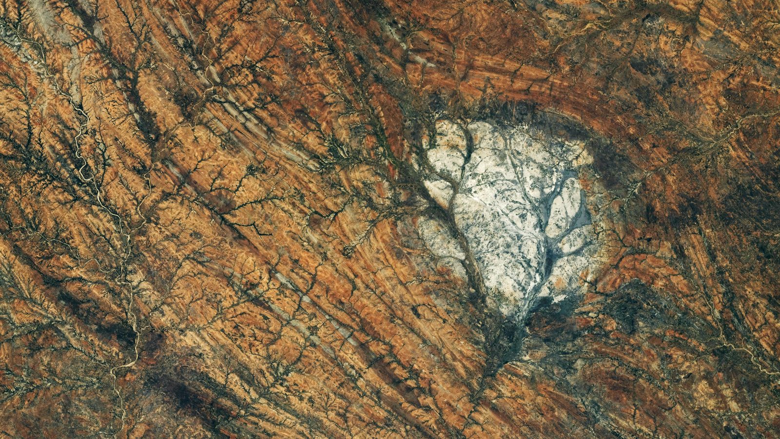 Mancha clara de anortosito em Madagascar vista da ISS, cercada por rochas laranja e marrons deformadas em faixas