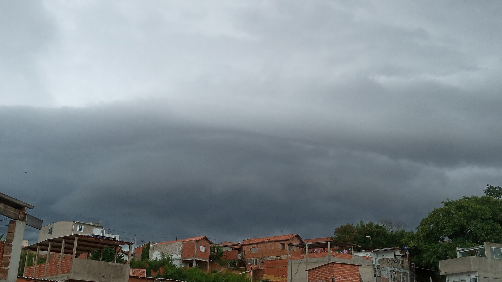 Nuvem escura em forma de rolo, uma frente de rajada, sobre casas de tijolo aparente e árvores, com o céu cinza ao fundo