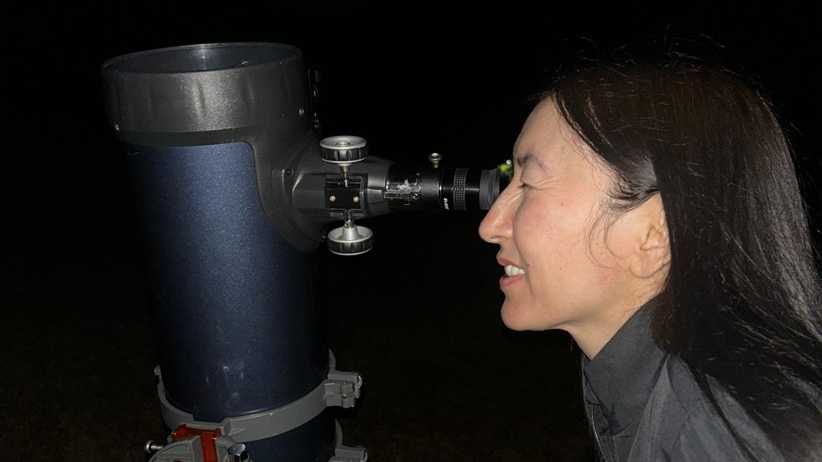 Mulher de perfil olhando por uma ocular lateral de telescópio refletor azul, à noite, numa observação dos anéis de Saturno
