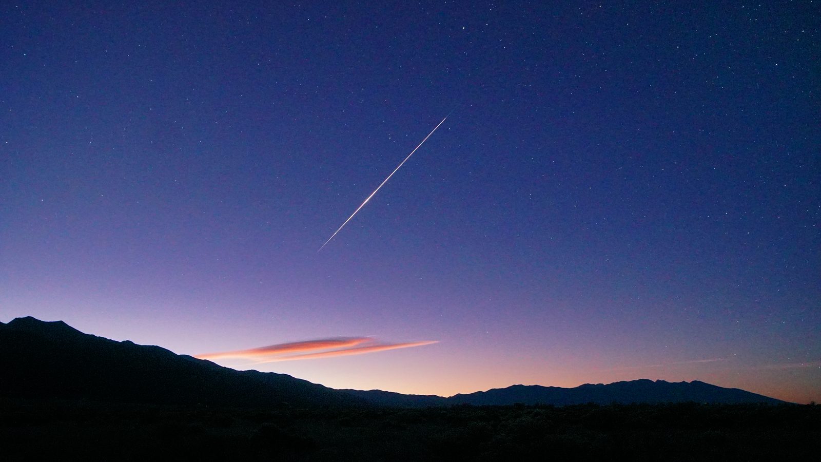 Meteoro de uma chuva de meteoros Orionídeas riscando o céu roxo do amanhecer sobre montanhas, com uma nuvem alaranjada perto do horizonte