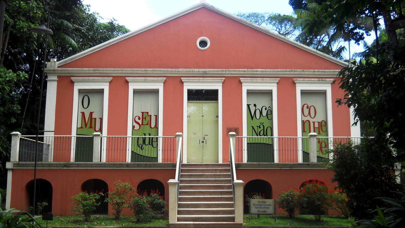 Pavilhão rosa com janelas cobertas por faixas no Parque Zoobotânico do Museu Goeldi, em Belém, cercado de árvores