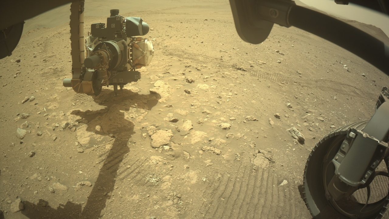Braço do rover Perseverance estendido sobre o solo de Marte, com as marcas das rodas e pedras claras ao fundo, no afloramento Idubi