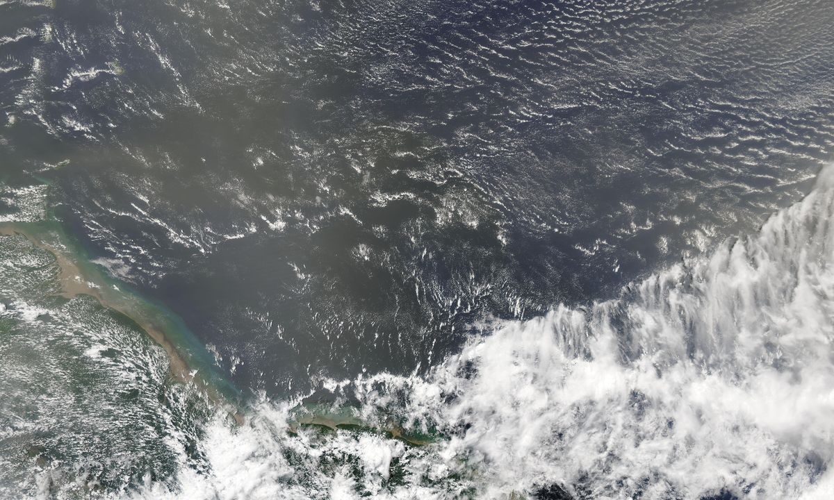 Oceano escuro com um véu bege de poeira sobre a água e a costa verde do norte da América do Sul no canto de baixo