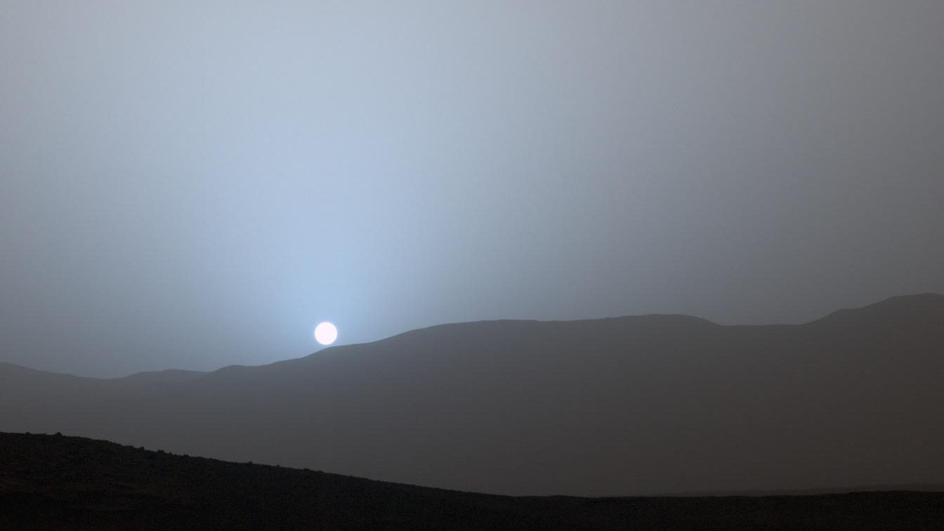 Pôr do sol azulado em Marte registrado pelo jipe Curiosity na cratera Gale, com o Sol baixo no horizonte e um halo azul em volta dele
