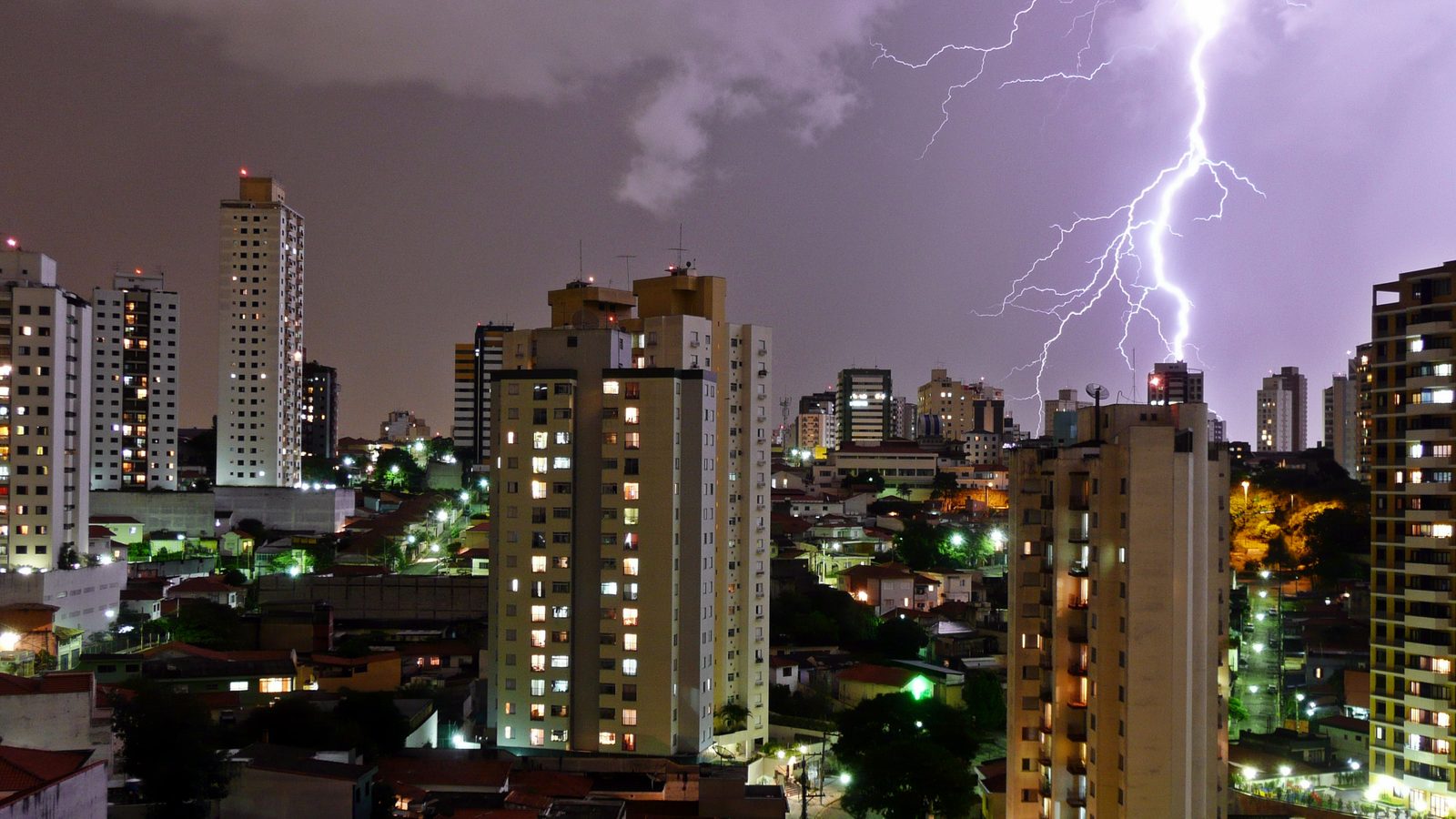 Raio cai sobre prédios e casas de São Paulo durante tempestade à noite, em céu roxo, cena típica dos avisos de tempestade do Inmet