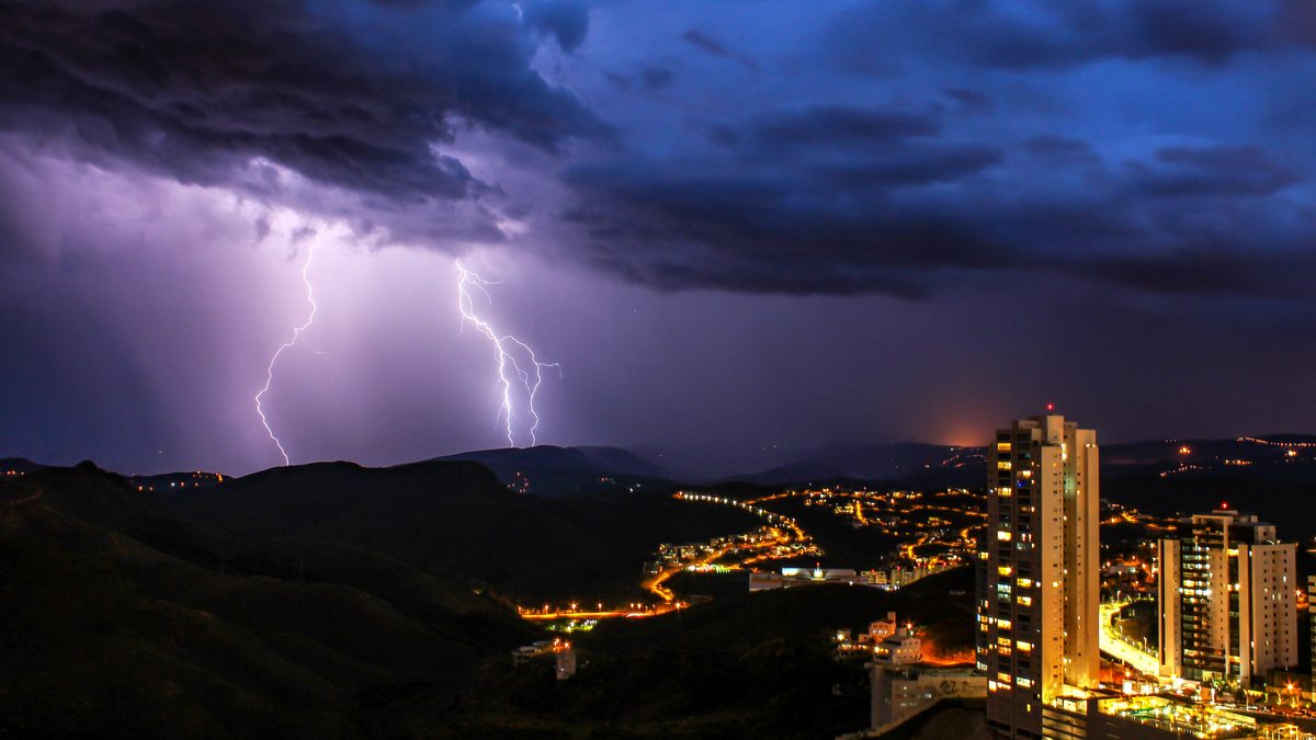 Raios caindo de nuvens escuras sobre os morros e prédios de Nova Lima à noite