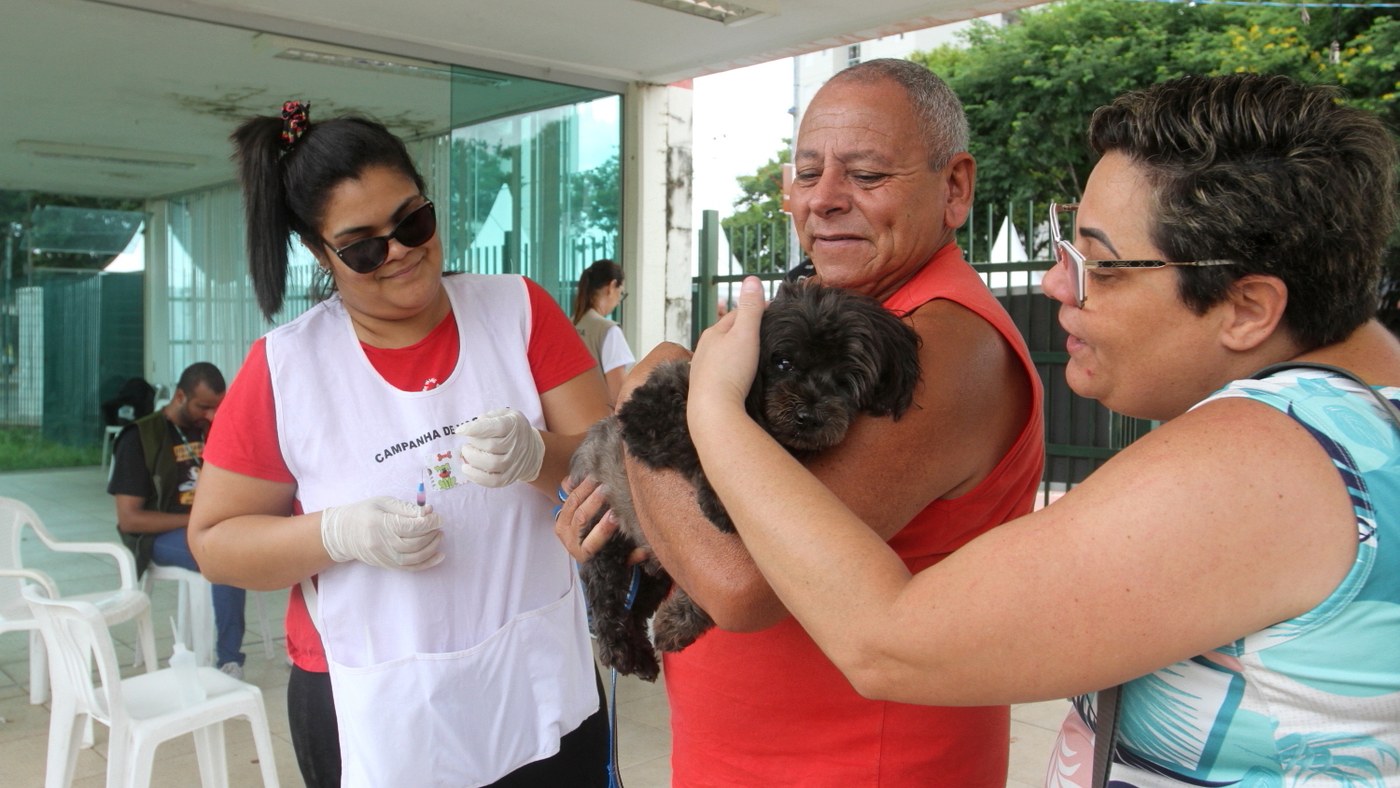 Dia Mundial contra a Raiva: em vacinação antirrábica da Prefeitura de Jacareí, agente de saúde de luvas se prepara para vacinar um cachorro preto no colo do dono