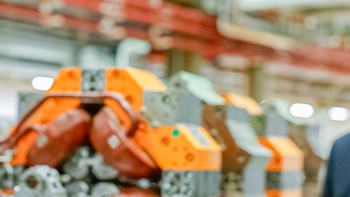 Peças laranja e cinza de um equipamento do Sirius em primeiro plano, num galpão com tubulações vermelhas ao fundo