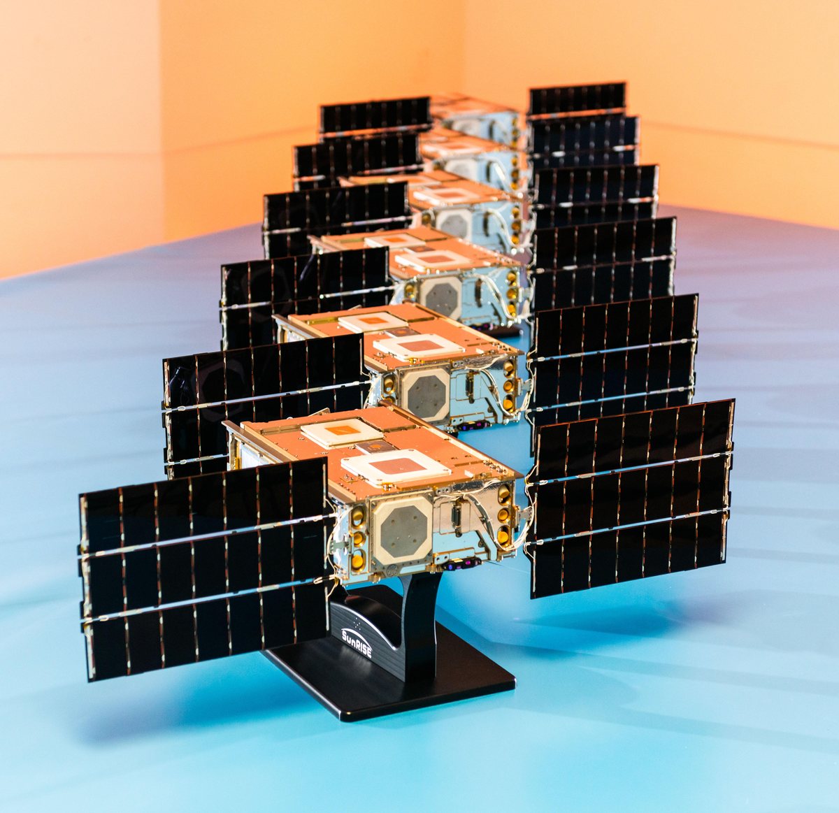 Os seis satélites da missão SunRISE em fila sobre uma mesa azul, o da frente num suporte preto com a inscrição SunRISE, contra fundo laranja